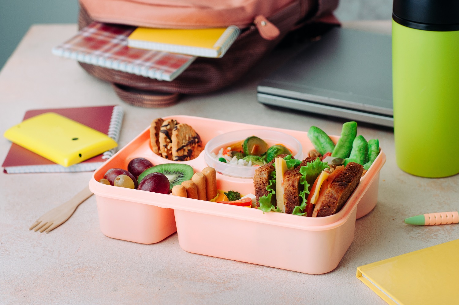 Open lunch box with healthy food on the table near backpack, lap